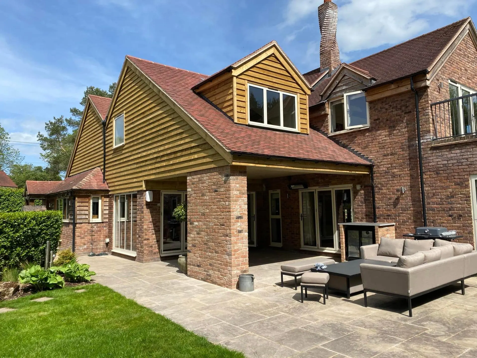 Rear elevation of Dingleside in Worcestershire showing a new two-bedroom extension with oak-clad gable and dormer, plain clay tile roof, brick columns and a covered outdoor entertainment terrace with bifold doors, built by JRC Developers