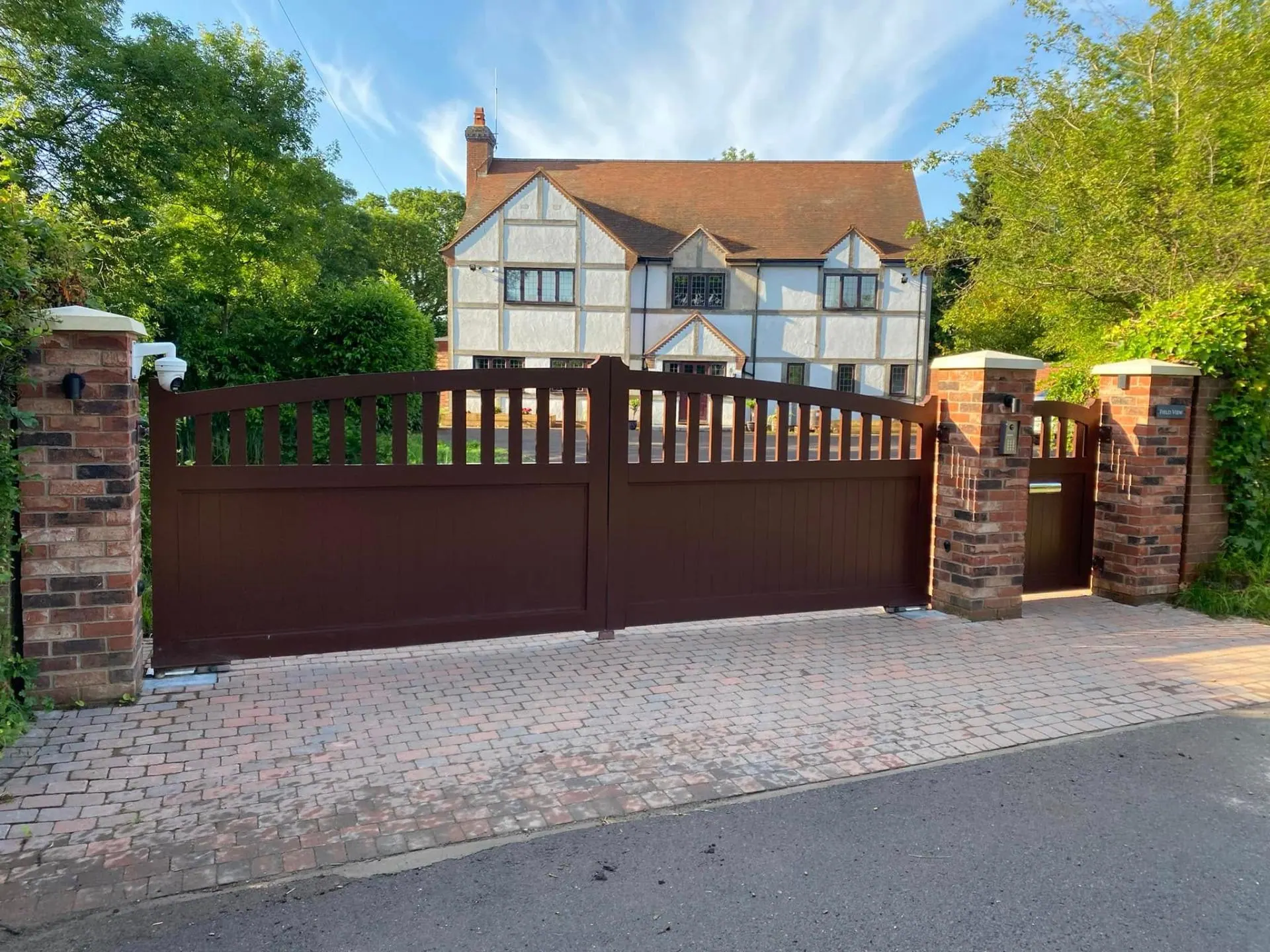 Automated timber driveway gates in dark oxblood finish with brick pillars, intercom and CCTV outside Field View, a Tudor-style home in Worcestershire built by JRC Developers