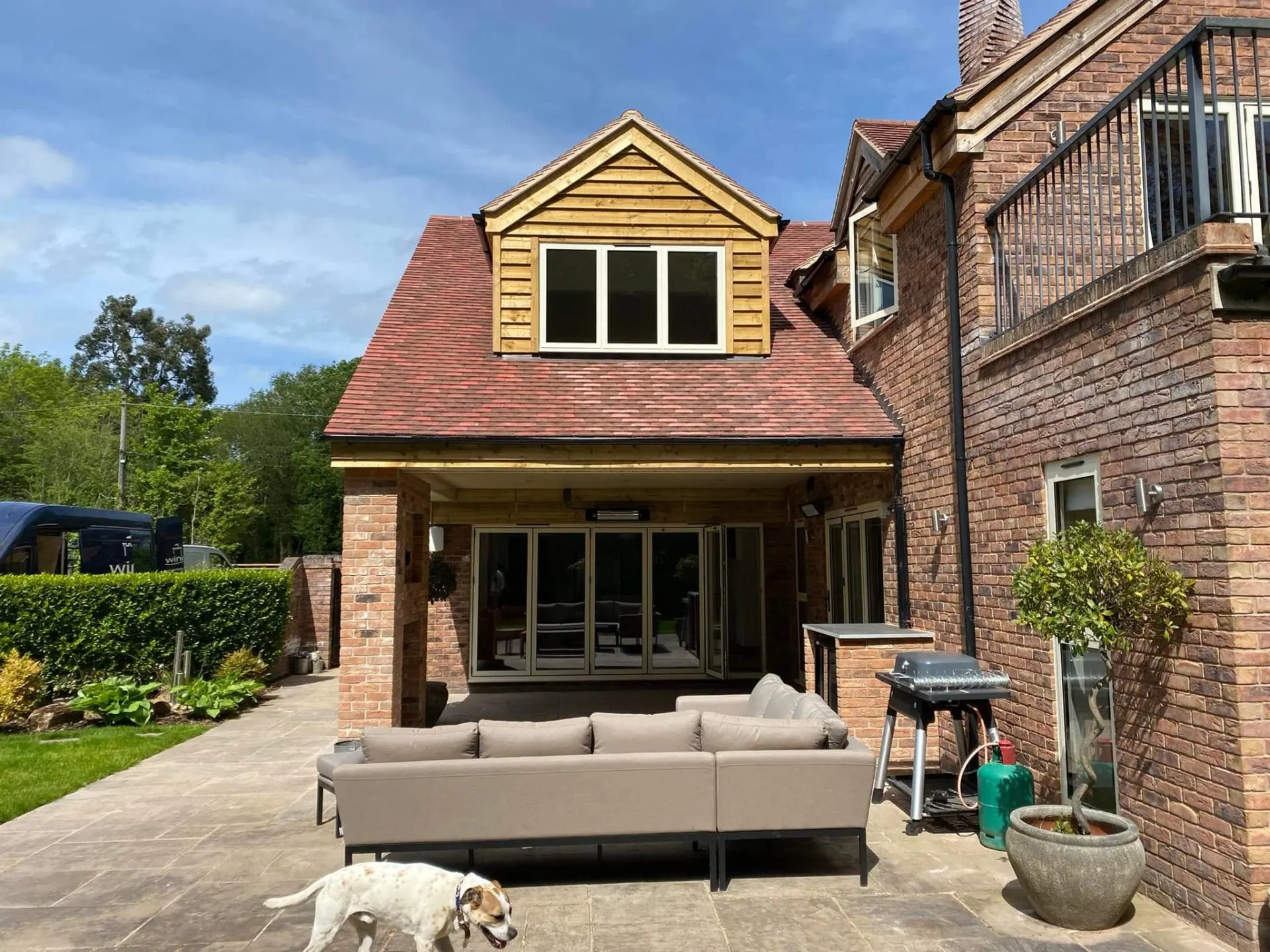 Front-on view of the covered outdoor entertainment area at Dingleside, showing the oak-clad dormer, plain clay tile roof, brick columns, aluminium bifold doors and Indian-stone patio with garden furniture, JRC Developers Worcestershire