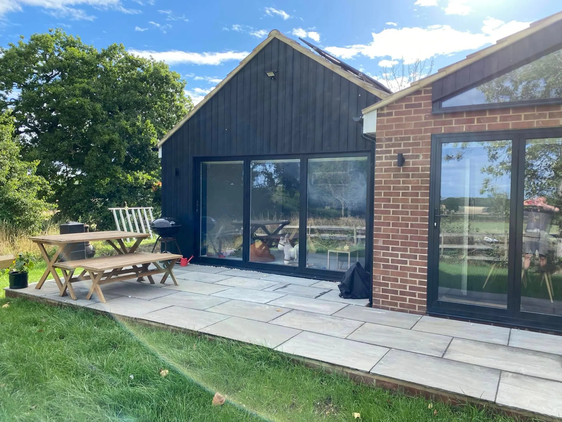 Black timber clad gable with wide aluminium sliding doors opening onto a sandstone patio with garden table and benches